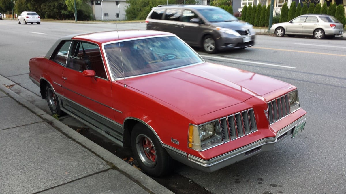 Photo of a red 1978 Pontiac Grand Am with color-coded wheels and a vinyl half-top, parked on a city street