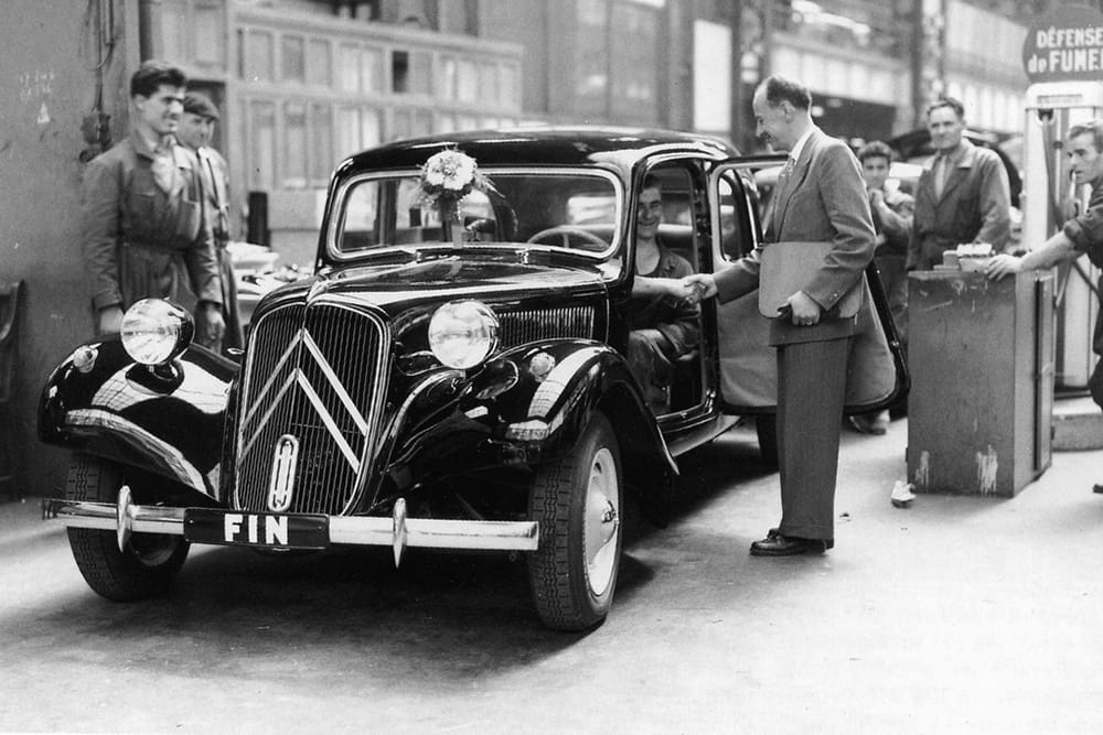 B&W photo of a black Citroen Traction Avant with a flower on the windshield and the word "FIN" on the front bumper coming out of a factory as smiling Frenchmen look on