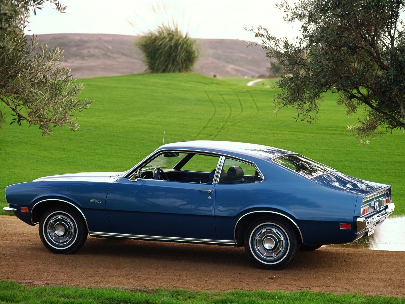 Press shot of the rear 3q of a blue 1970 Ford Maverick on a country road