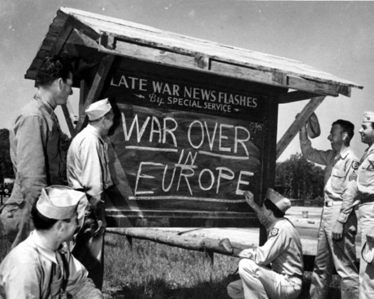 Posed shot of U.S. servicemen standing around an outdoor chalkboard on which one is writing "Late War News Flashes By Special Service: War Over in Europe"
