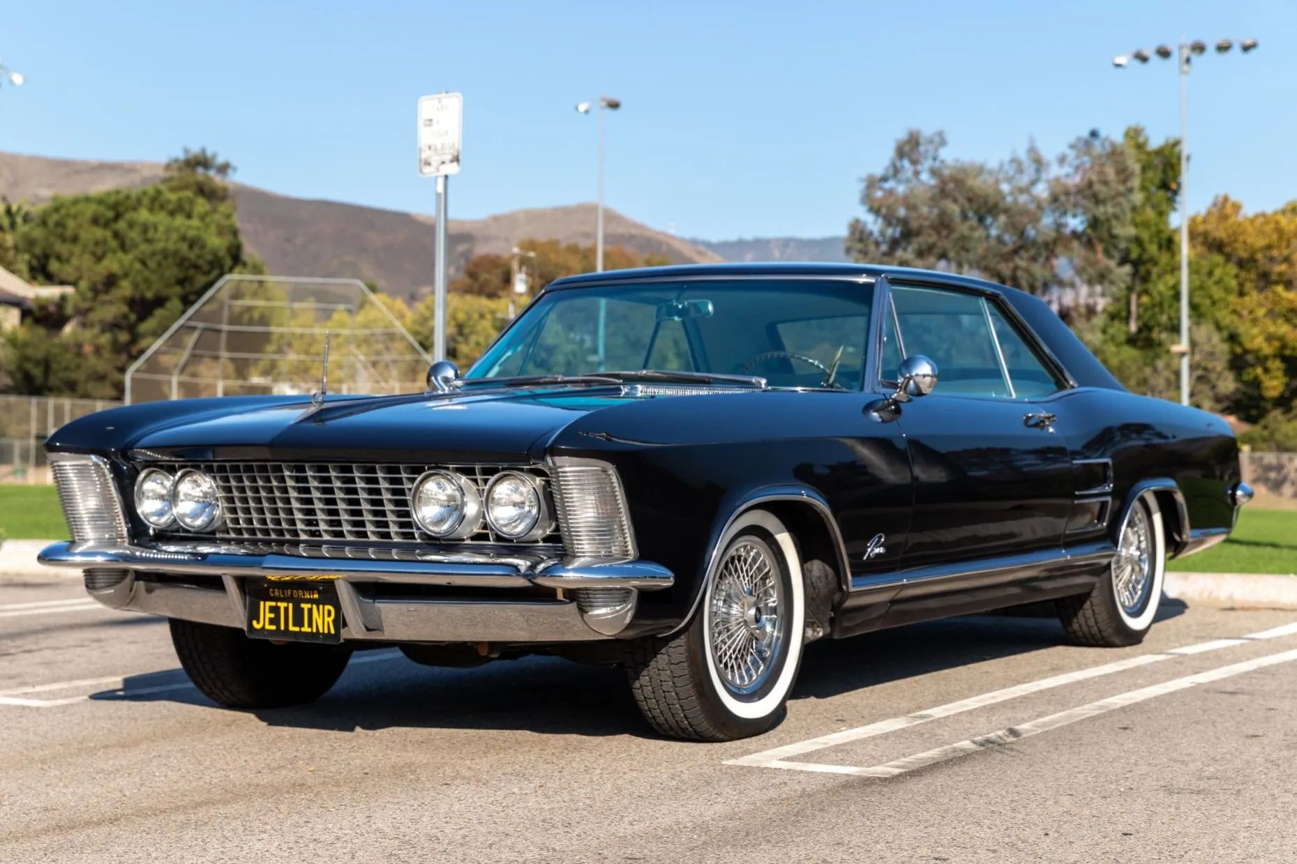 Front 3q view of a Diplomat Blue 1964 Buick Riviera with black and yellow license plate reading "JETLINR," wire wheel covers, and whitewall tires, in the parking lot of a park