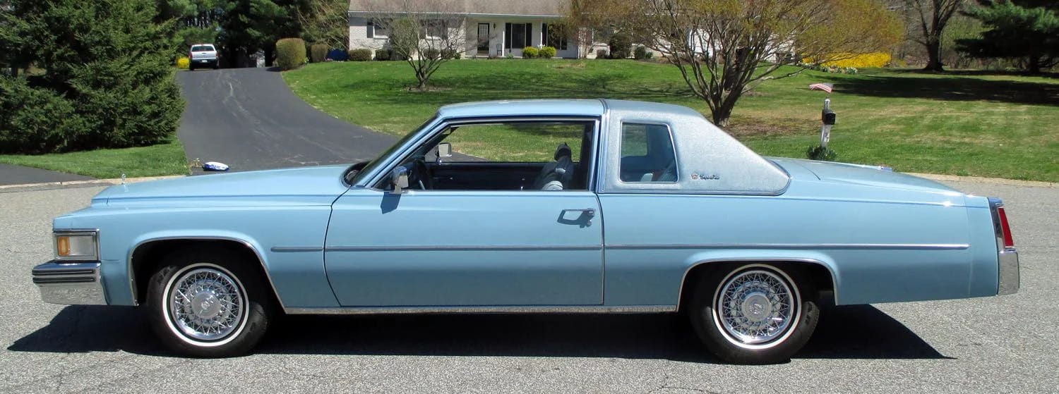 Left side view of a light blue 1977 Cadillac Coupe de Ville with a blue vinyl top and wire wheel covers