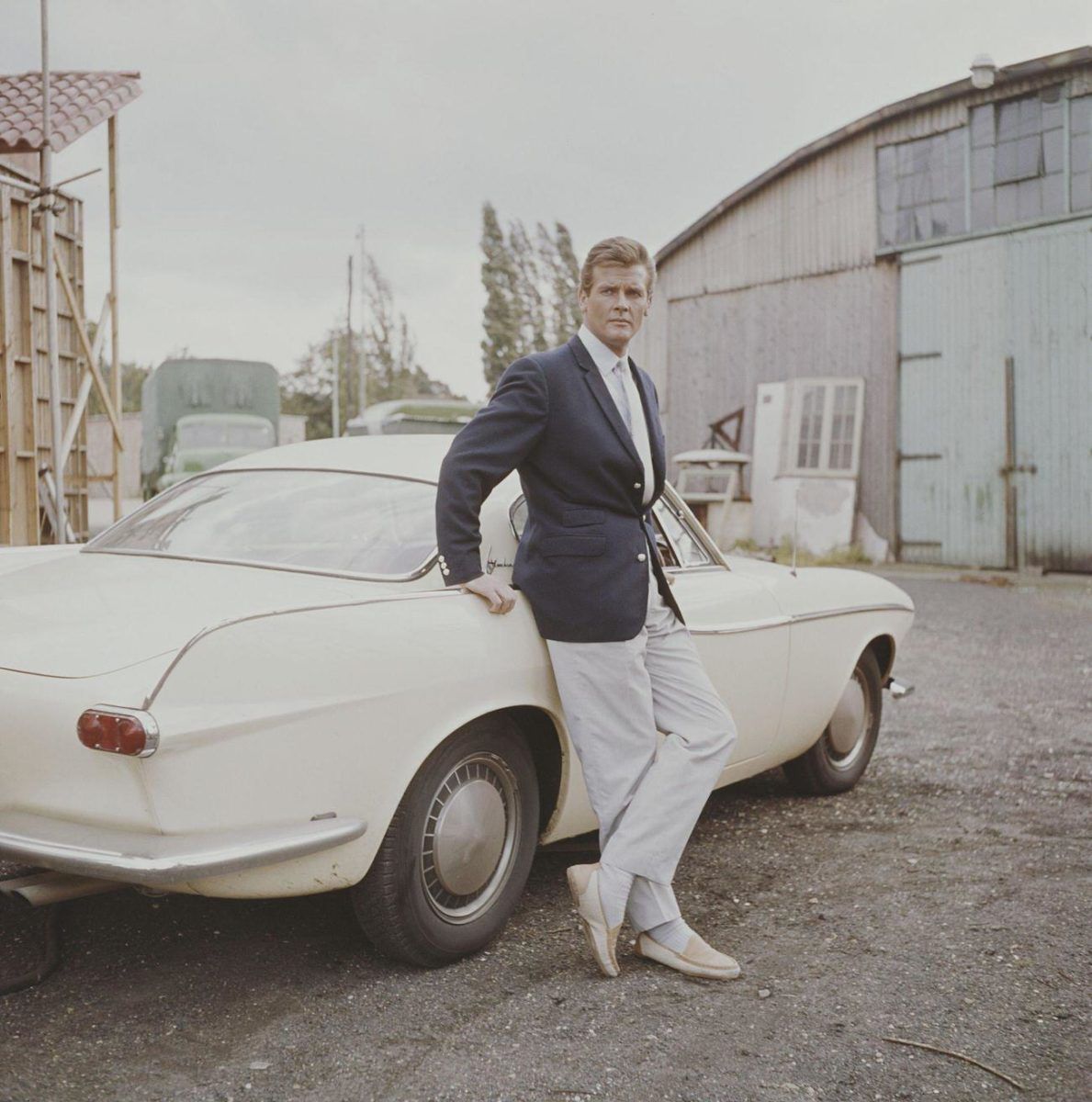 Old color photo of Roger Moore leaning against a white Volvo 1800S