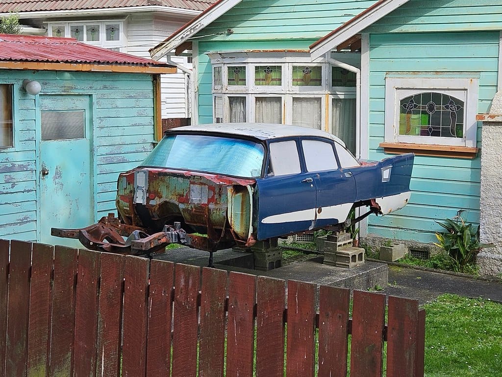 Image of a 1957-1958 Plymouth sedan with its front end missing, displayed on concrete blocks in a house's front yard. 