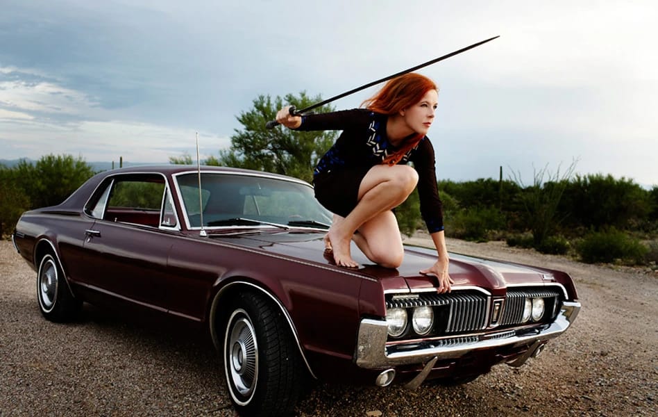 Photo of singer Neko Case, holding a sword, crouching on the hood of a burgundy 1967 Mercury Cougar with a black vinyl top