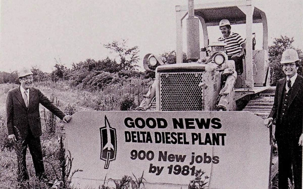 1985 Cadillac De Ville 4.3 V-6 Diesel: The Most Underpowered Cadillac Since Before WW2 8 B&W photo of two smiling white men in suits and hard hats standing next to a bulldozer whose blade is labeled "GOOD NEWS Delta Diesel Plant: 900 new Jobs by 1981" with the Oldsmobile logo