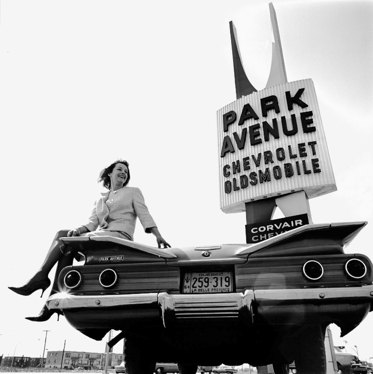 A low angle rear view of a 1960 Chevrolet, with a model sitting on the trunk, and a modernist-era sign of the Park Avenue Chevrolet dealer in Quebec. 