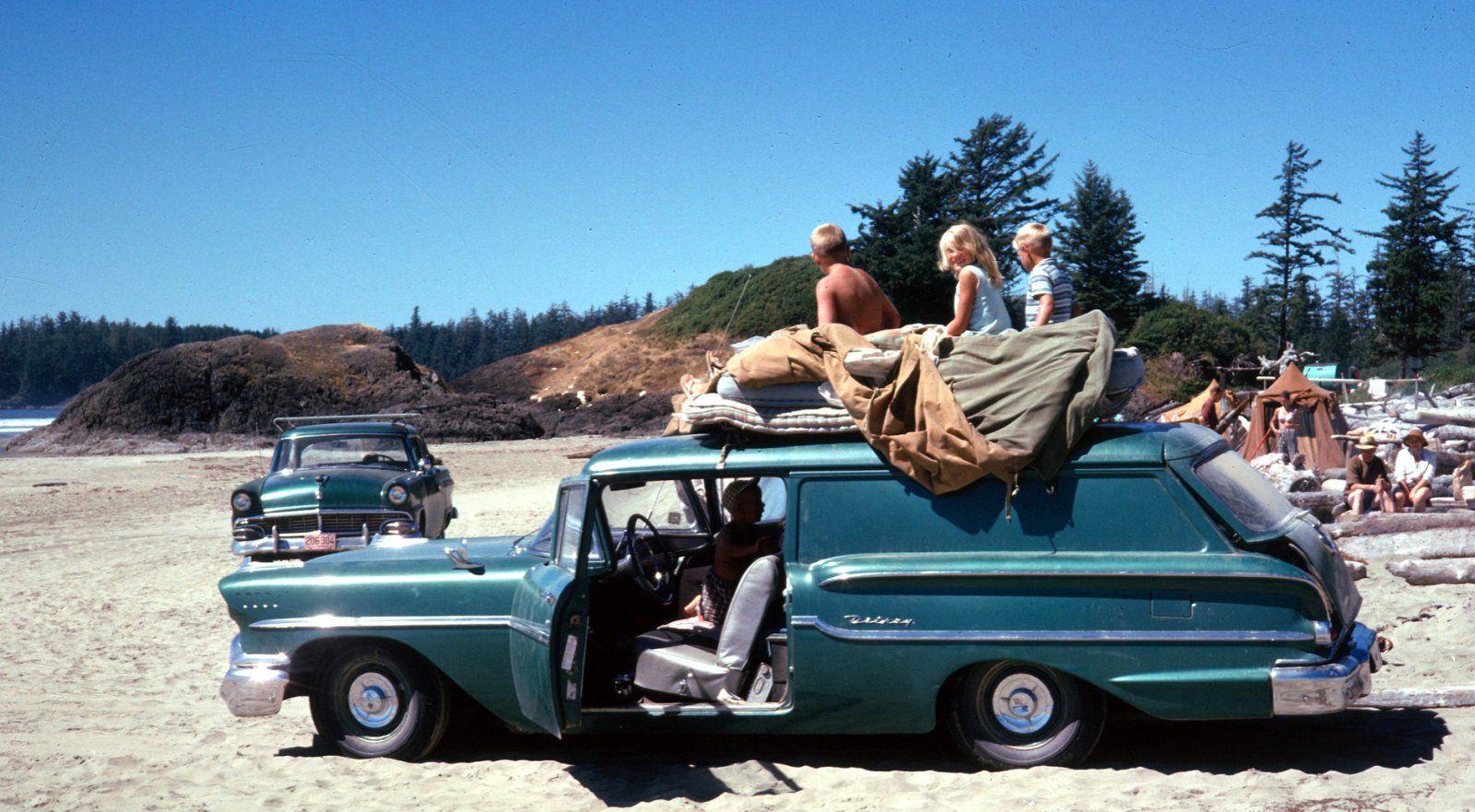 Vintage photo of three young children sitting on camping gear on the roof of a green 1958 Chevrolet Delray sedan delivery, parked on sand