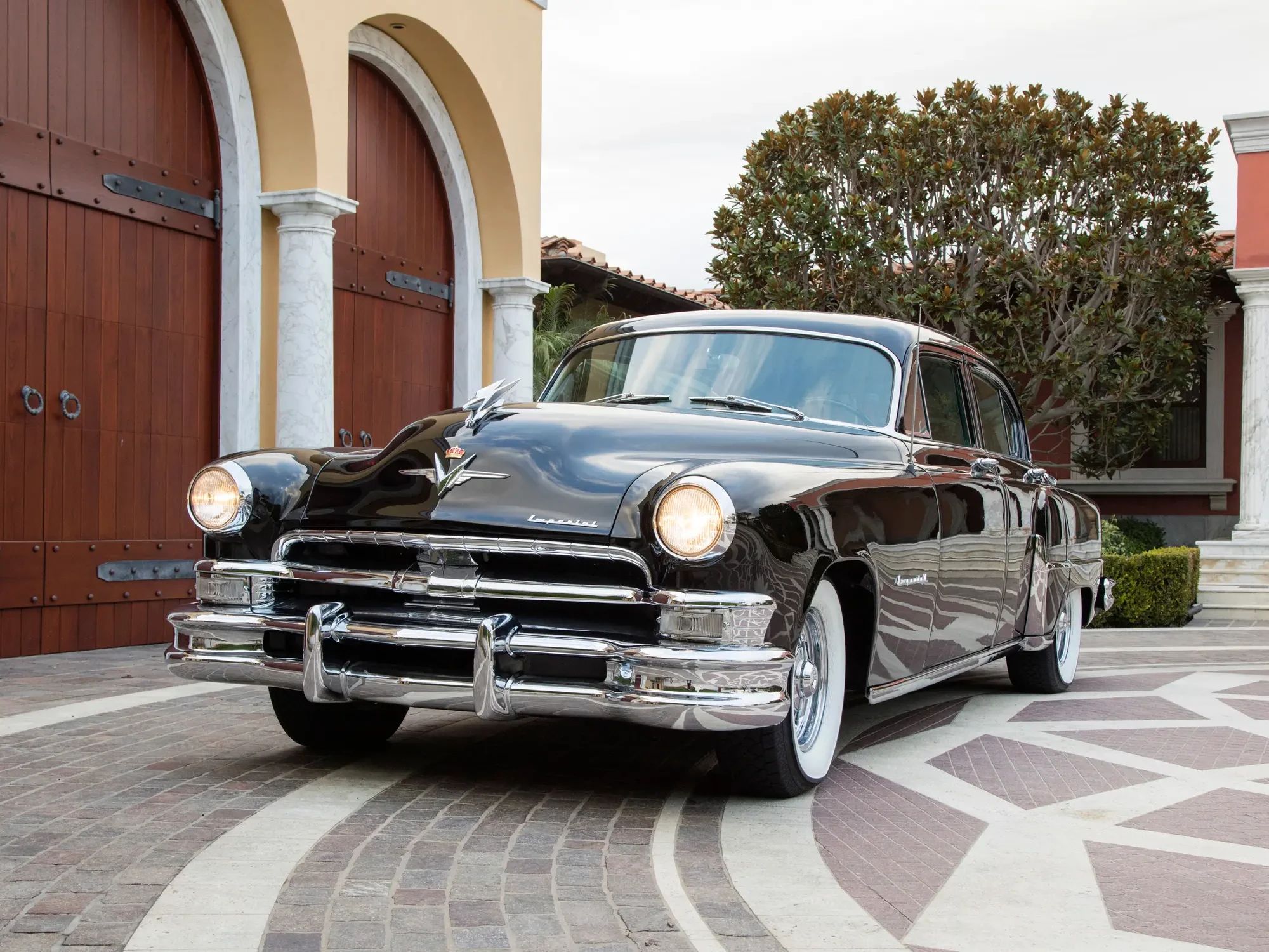 Left front 3q view of a black 1953 Chrysler Custom Imperial with its headlights on, parked next to an archway
