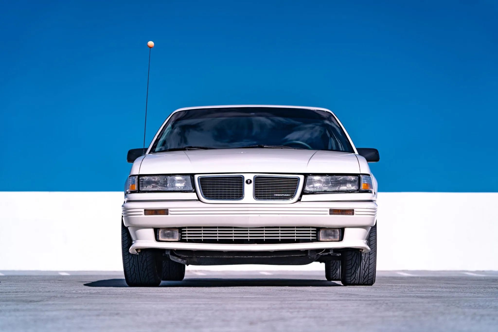 Low-angle front view of a white 1990 Pontiac Grand Am SE framed against a bright blue sky