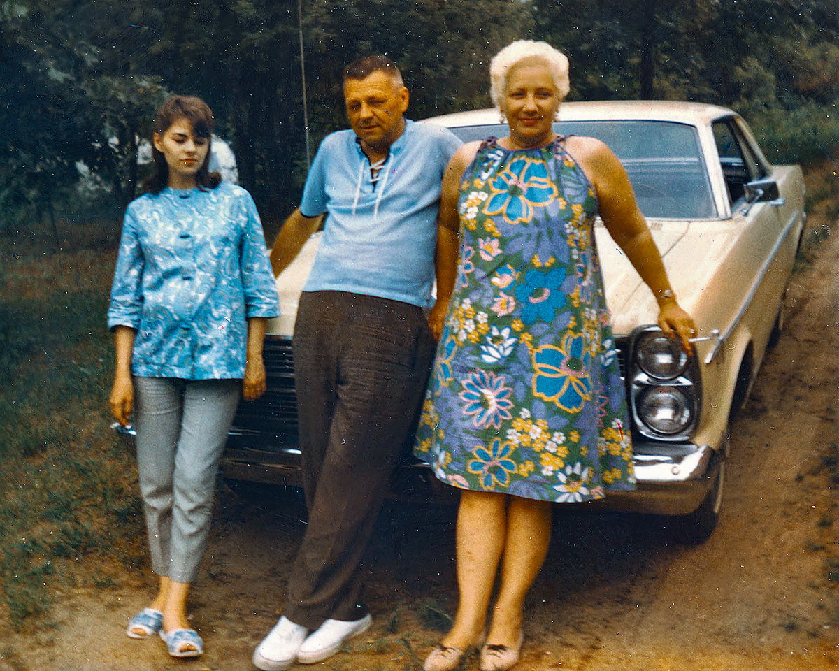 Vintage color photo of a young woman standing next to an older couple leaning on the hood of a yellow 1966 Ford Galaxie