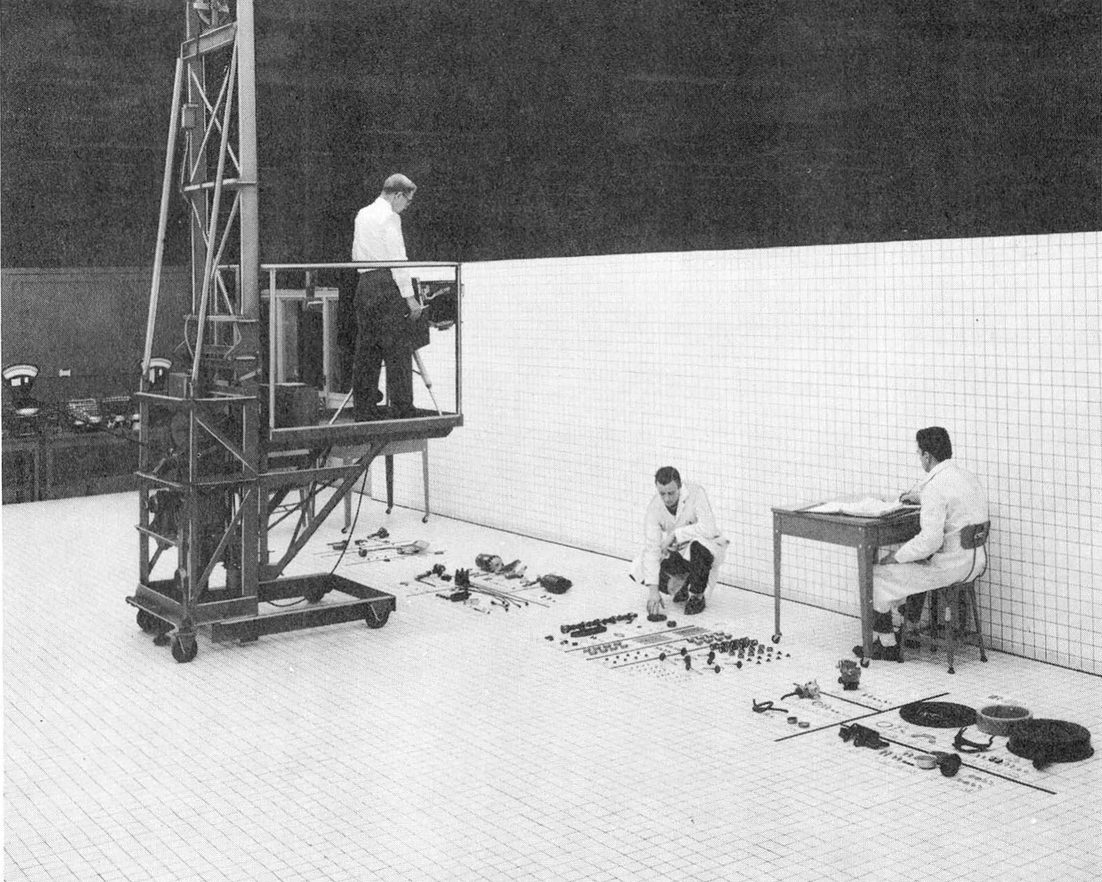 B&W photo of a Ford engineer on a scaffold as two other engineers lay out parts on mats on the floor