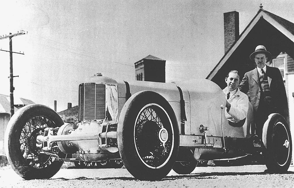 B&W photo of a low-slung FWD race car with a clean-shaven white man at the wheel and a white man with a hat and mustache standing next to him
