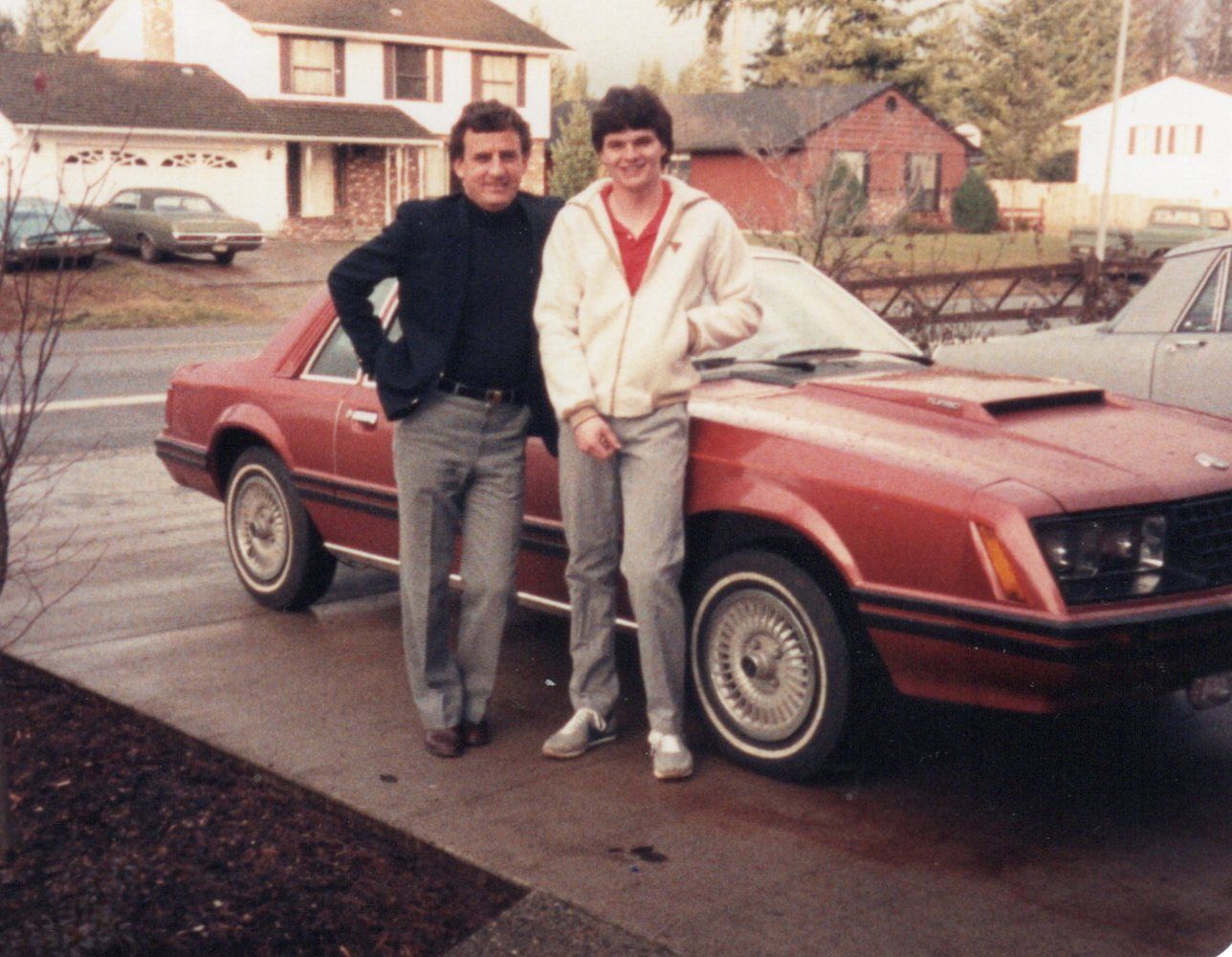 Photo of a man and a boy posing in front of a red 1980 Ford Mustang
