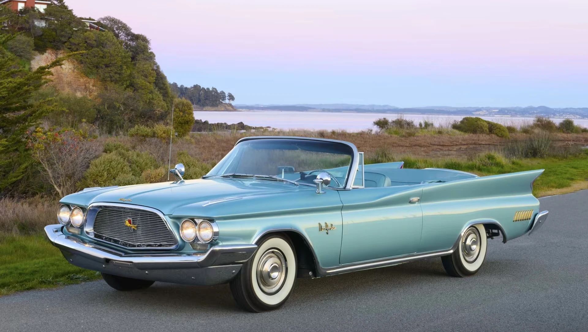 Photo of a turquoise 1960 Chrysler New Yorker convertible parked near a beach with a dramatic purple-and-blue sunset sky