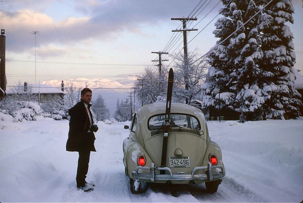 Photo of a man standing in a snowy road next to a VW Beetle with skis strapped to the back