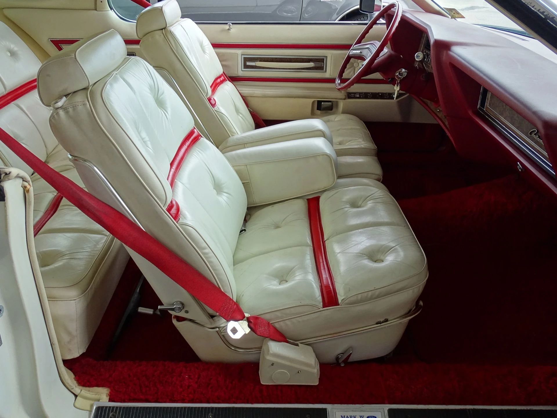 Front seat of a 1975 Lincoln Mark IV with red accents on white leather seats and bright red seatbelts