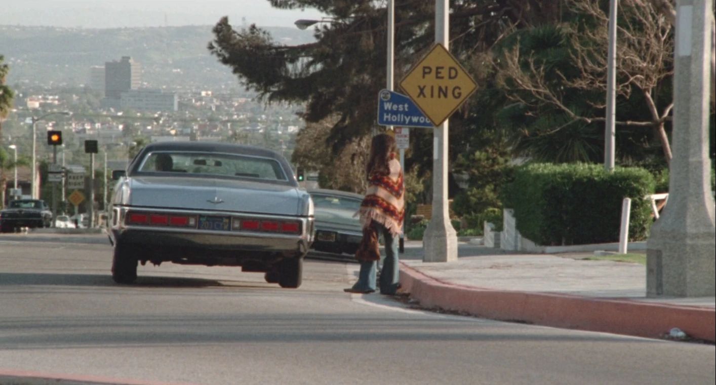Why The 1972 Lincoln Continental Was The Perfect Movie Costar In This Jack Lemmon Drama 16 Movie screenshot of a gray 1972 Lincoln Continental sedan pulling away from the curb on a Los Angeles street, with Laurie Heineman standing at the curbside under signs reading "West Hollywood" and "PED XING"