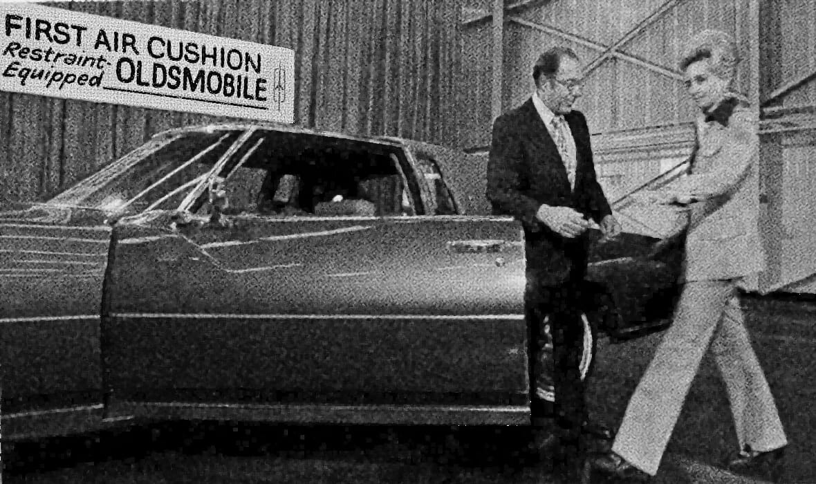 Photo of a man standing in the door of a 1974 Oldsmobile Toronado beneath a banner reading "FIRST AIR CUSHION Restraint-Equipped OLDSMOBILE"