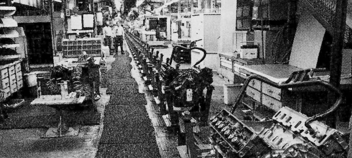 B&W photo of men watching an engine come down an empty assembly line