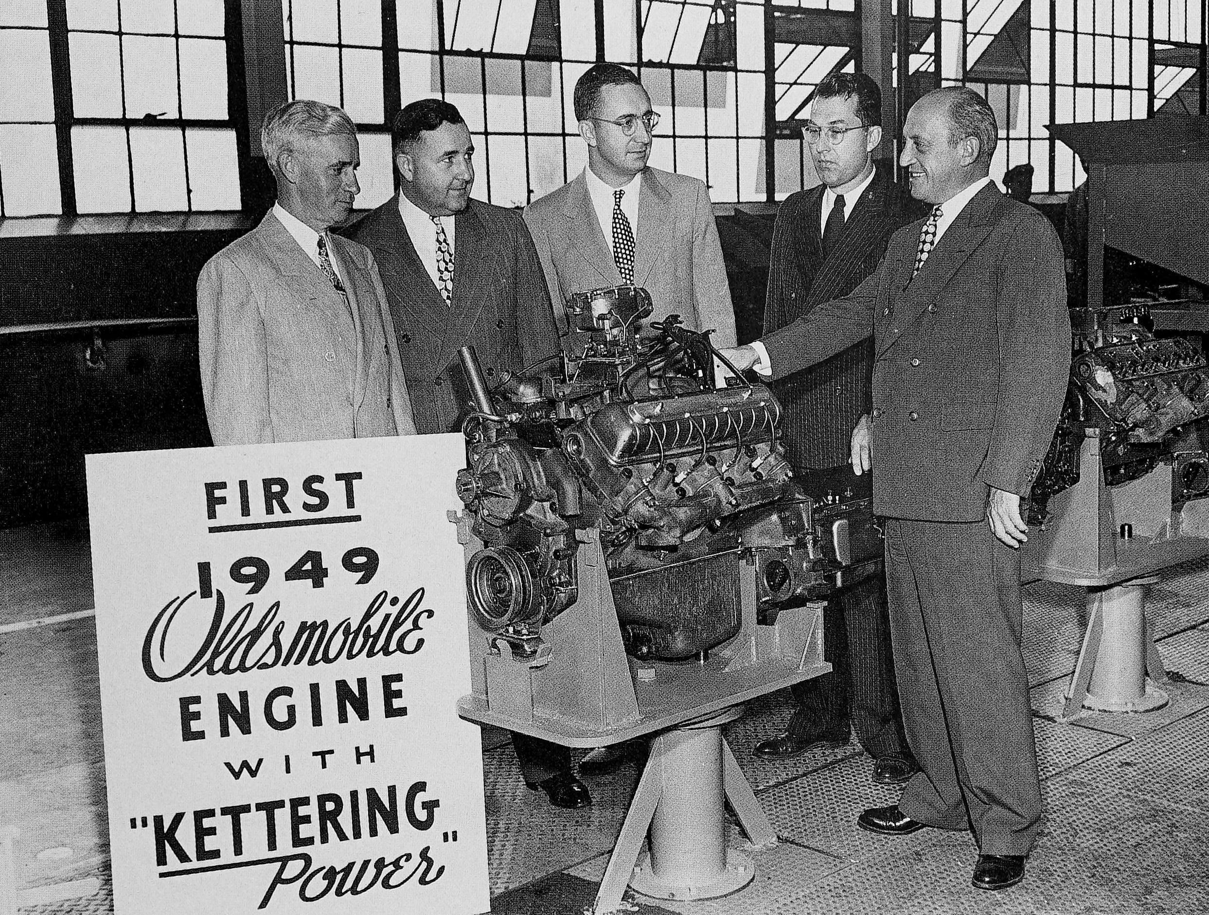 1949 Oldsmobile Rocket V-8 And The Ultra-High-Compression Economy Version That Never Made It 10 B&W photo of five men in suits standing around a display engine with a sign reading 'First 1949 Oldsmobile engine with 'Kettering Power'"