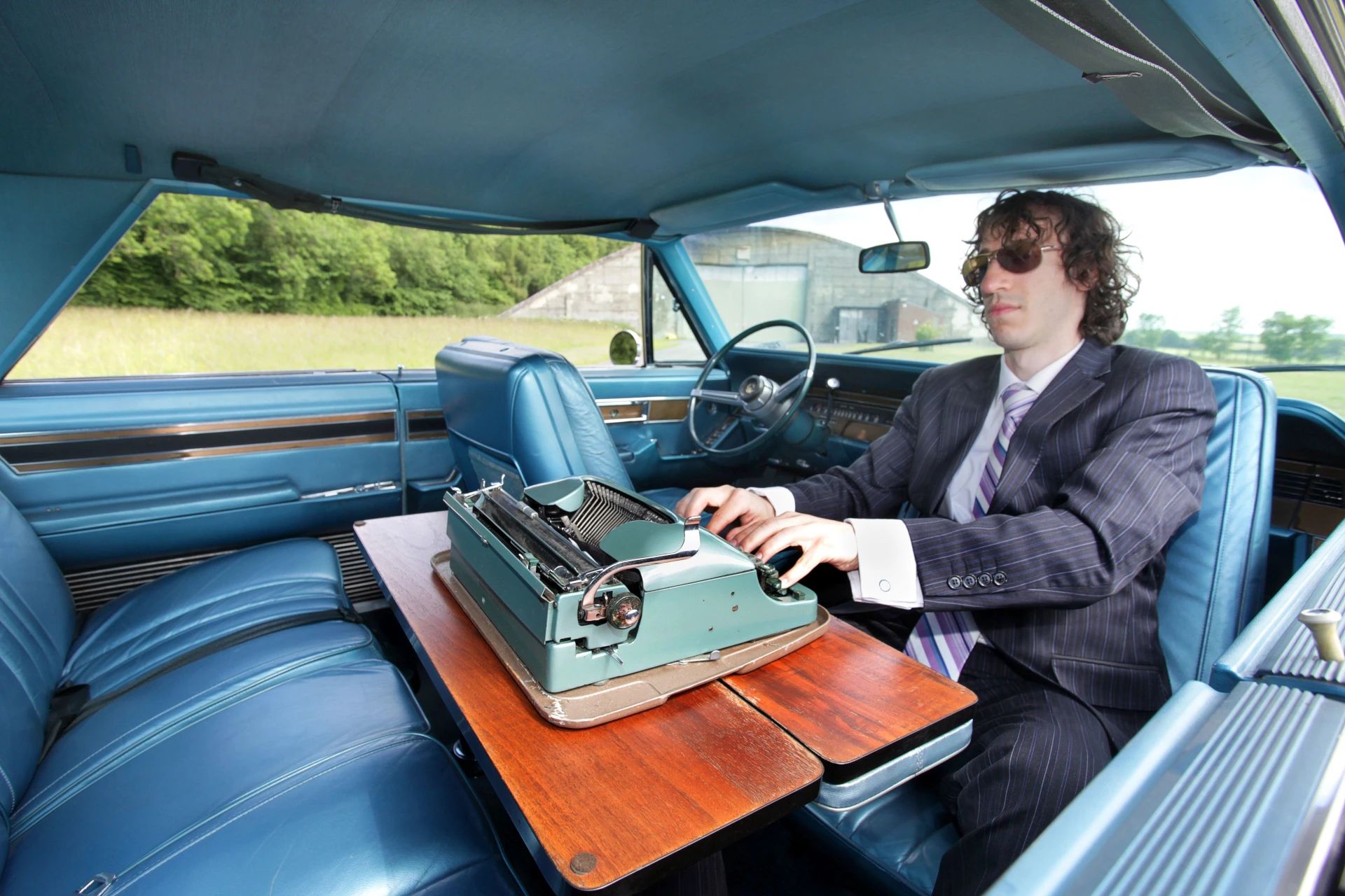 Interior of a 1968 Imperial with a folding desk and front swivel seat; a young man in a suit and sunglasses in the right front seat is pretending to type on a mechanical typewriter