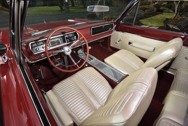 Dashboard and front bucket seats of a Bright Red 1967 Plymouth GTX with white vinyl upholstery and center console, viewed through the driver's door