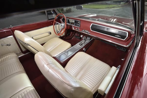 Dashboard and front bucket seats of a Bright Red 1967 Plymouth GTX with white vinyl upholstery and center console, viewed through the passenger door
