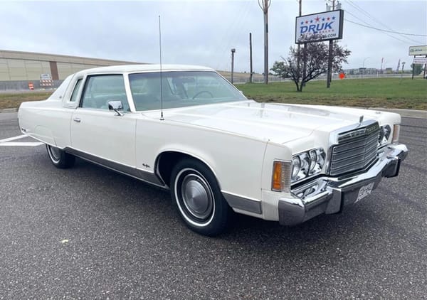 Front 3q view of a white 1974 Chrysler New Yorker Brougham two-door hardtop; there's a Druk Auto Sales sign in the background